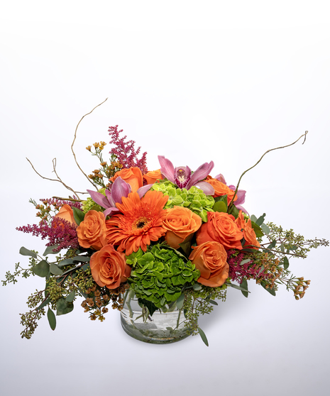 green hydrangea, orange roses, orange gerbera daisies, cymbidium orchids and pink astilbe, accented by curly willow and seeded eucalyptus greenery in an off white vase